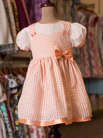 A sweet peach-colored gingham dress with puffy white sleeves. This classic pinafore style is timeless, comfortable, and perfect for a picnic or playdate.