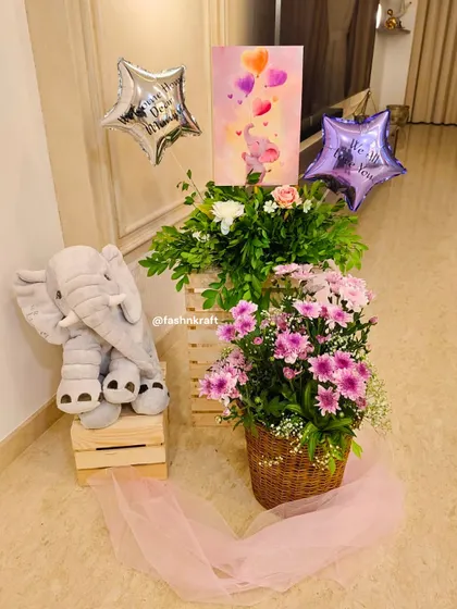 A sweet corner setup for a baby girl's arrival. It features a plush elephant toy, star-shaped balloons, and baskets of fresh pink and white flowers, creating a soft and celebratory welcome.