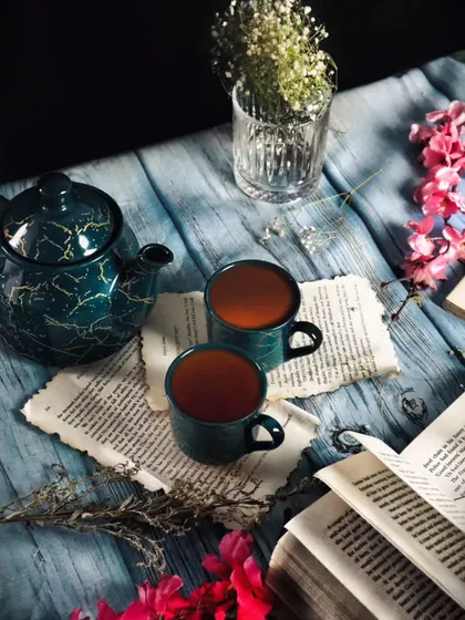 A moody flatlay with two cups of tea, a beautiful teapot, an open book, and flowers on a blue wooden surface. The composition and colors create a rich, aesthetic, and calming scene.
