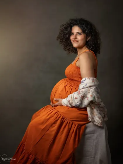 A warm and gentle studio portrait of a mother-to-be in a comfortable orange dress, her curly hair framing a soft smile. The simple, classic backdrop keeps the focus entirely on her serene expression.