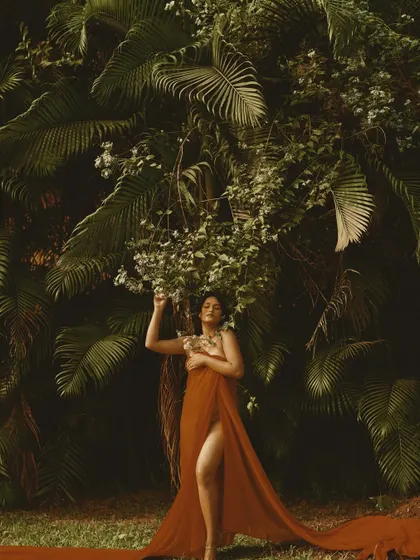 A wide shot that shows the full effect of the flowing red fabric against the lush green background. The hair and makeup are designed to feel organic and part of the natural world.