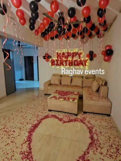 A romantic birthday surprise with red and black balloons covering the ceiling. The setup includes a 'Happy Birthday' foil balloon banner and a heart made of rose petals on the floor.