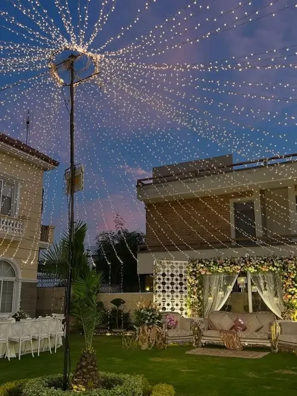 A beautiful outdoor evening setup with a canopy of fairy lights stretching across the lawn. The stage in the background is elegantly lit, creating a magical atmosphere.
