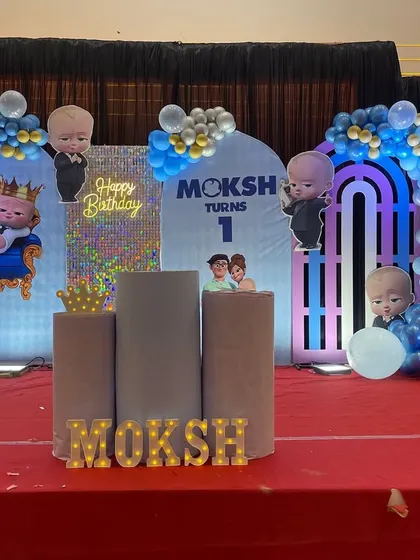 A large-scale 'Boss Baby' themed first birthday party on a stage. This setup includes multiple character cutouts, custom backdrops, marquee letters spelling the baby's name, and extensive balloon work in shades of blue and silver.