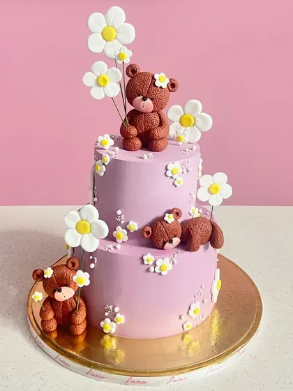 A sweet and charming two-tiered cake featuring adorable teddy bears and delicate fondant daisies. The lavender color and playful design make it perfect for a first birthday or baby shower.