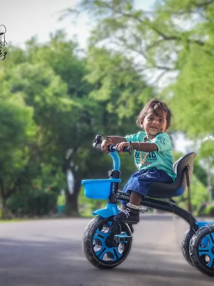 An action shot of a little boy enjoying his tricycle outdoors. The photo is taken on a quiet road lined with trees, capturing a classic childhood moment full of energy.