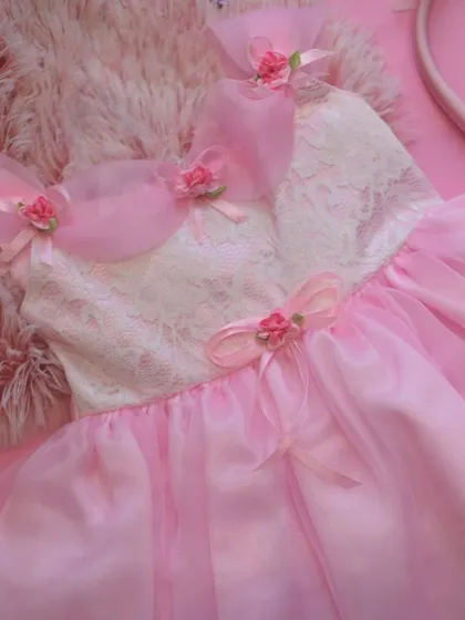 A fairytale dress in soft pink. This design features a delicate white lace bodice and layers of sheer pink fabric, adorned with tiny roses and bows for an incredibly sweet and romantic look.