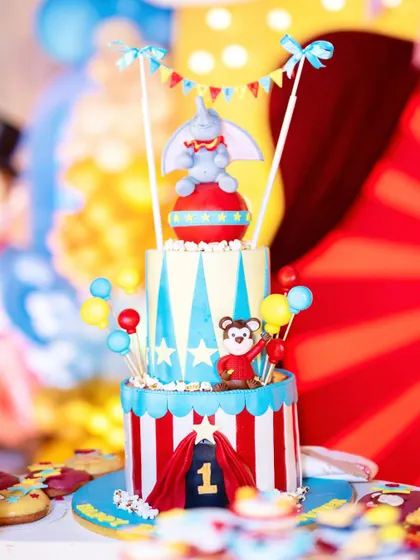 A whimsical, multi-tiered cake for a carnival-themed first birthday. The cake is adorned with a cute Dumbo topper, a monkey, and other circus elements in bright, cheerful colors.