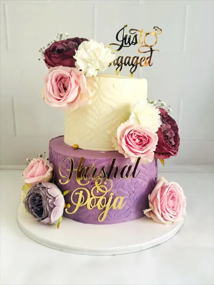 A beautiful two-tier engagement cake with contrasting textures. The top tier is smooth and white, while the bottom features a purple textured design, all brought together with fresh flowers.