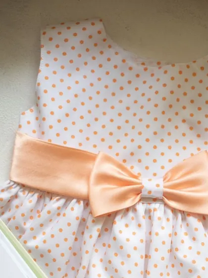 A sweet and simple design in white with orange polka dots. The dress is cinched with a wide peach satin sash that ties into a beautiful bow, adding a soft touch of color.