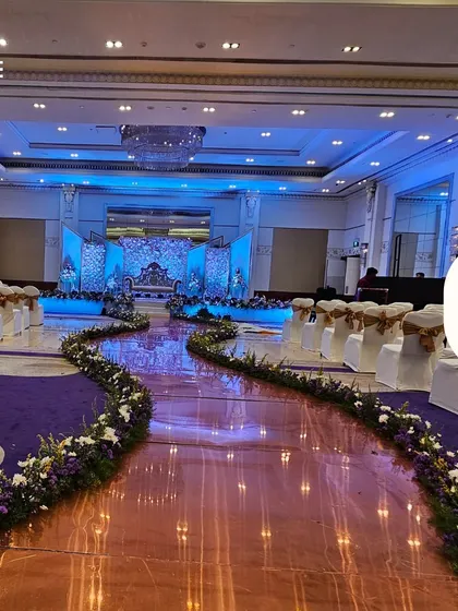 A wide view of a reception hall with a reflective purple aisle leading to the main stage. The aisle is lined with floral arrangements, guiding guests toward the beautifully lit blue and white stage.