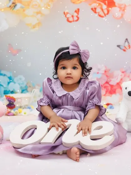 A sweet portrait of a one-year-old girl in a purple dress, holding a "one" sign in a whimsical, colorful setup.