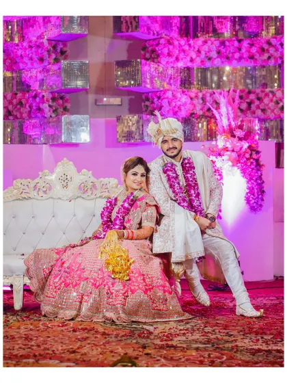 A formal portrait of the couple seated on an elegant white sofa against a backdrop of pink lighting and floral decor. This is a classic stage shot from an Indian wedding.