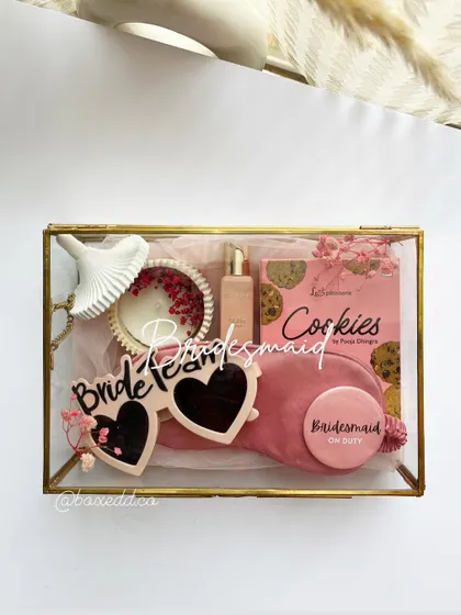 A chic and modern bridesmaid proposal in a glass box. This hamper features 'Bride Team' sunglasses, a velvet eye mask, a personalized badge, and other pink-themed goodies for a fun and aesthetic gift.