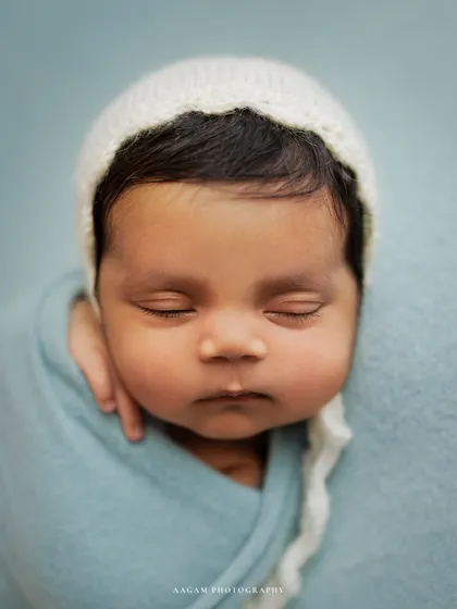 Those squishy cheeks are just irresistible. A close-up portrait of a newborn in a simple white bonnet against a soft blue background.