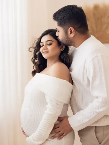 A sweet and protective gesture. The father-to-be kisses his partner's forehead while cradling her and the baby bump, a pose that radiates warmth and care.