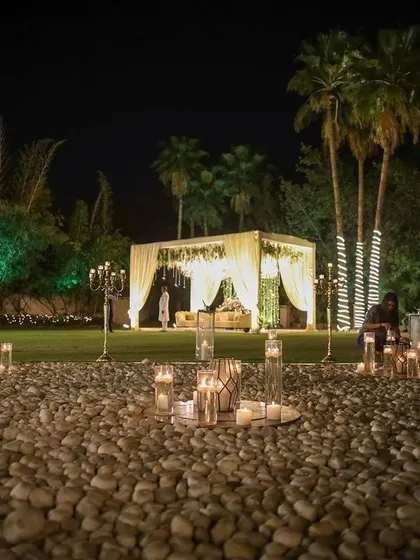A serene and romantic corner of an outdoor venue. A path of white pebbles is lined with candles in glass holders, leading to a beautifully draped mandap in the distance.