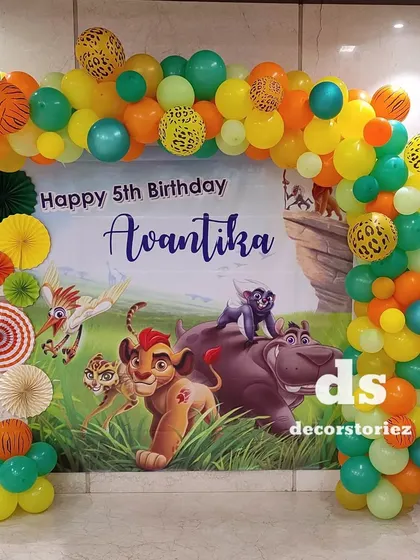 A Lion Guard themed 5th birthday party backdrop. The personalized banner featuring Simba and friends is framed by a colorful balloon arch with animal-print and paper fan details.