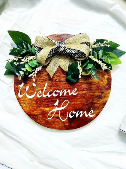 Our bestselling "Welcome Home" board. This circular wooden sign is decorated with a rustic bow and artificial leaves, creating a warm and inviting entryway piece.