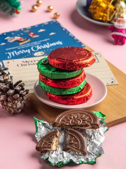 A stack of our chocolate-coated biscuits, wrapped in festive red and green foil. This image shows one unwrapped to reveal the crunchy biscuit and chocolate coating inside.