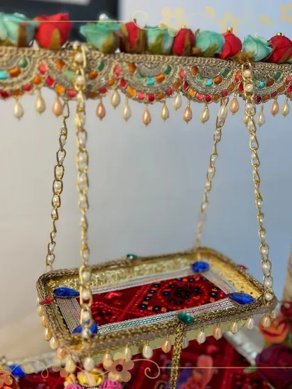 A detailed shot of the swinging seat of our Krishna jhula. The golden chains, rich red fabric, and pearl-drop border showcase the luxurious and divine quality of our handcrafted decor.