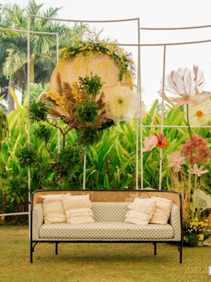 A cozy seating nook from the 'Golden Origami Garden' event. A modern cane sofa is set against a backdrop of lush greenery and giant, whimsical paper flowers, creating a perfect photo opportunity.