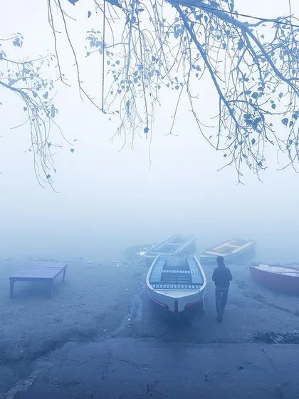A lone figure walks past empty boats on the shore of the Yamuna, the entire scene enveloped in a thick, atmospheric fog.