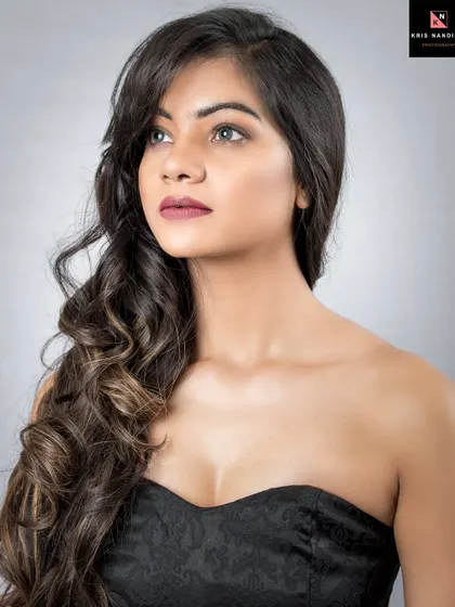 A classic beauty portrait against a simple grey studio background. The model wears a strapless black dress, and her hair is styled in soft waves, creating a timeless and elegant look.