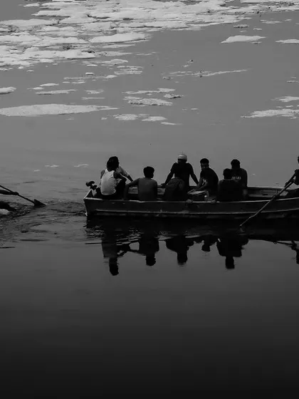 A black and white shot of people in a boat on the polluted Yamuna, their reflections stark against the water. The image is a somber look at the river's condition.