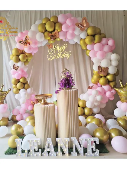 A royal-themed birthday decoration with pink, white, and gold balloons, crown props, and butterfly accents.