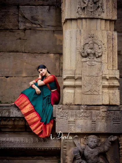 A serene pose in a red and green half-saree, seated against the detailed carvings of a temple wall. This shot captures the quiet elegance of the traditional attire.