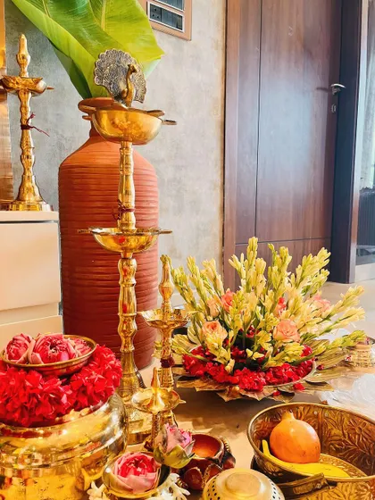 A collection of traditional decorative items, including ornate brass lamps (kuthu vilakku), a floral arrangement of tuberoses, and bowls of fresh flowers, creating a rich and auspicious display.
