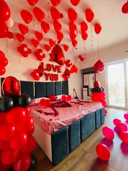 An intense red and black romantic room decoration. The setup includes "I Love You" foil balloons, heart-shaped balloons, and rose petals on the bed.