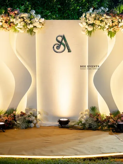 A modern and minimalist Sangeet backdrop. The design features clean, curving lines with integrated backlighting and a custom couple's monogram, topped with lush floral arrangements for a touch of organic elegance.