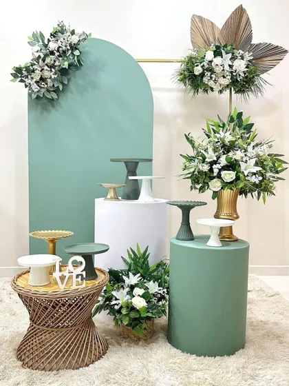 A sophisticated "no balloon" setup. This design uses a sage green arch, plinths, and beautiful white and green floral arrangements for an elegant and minimalist photo booth or backdrop.