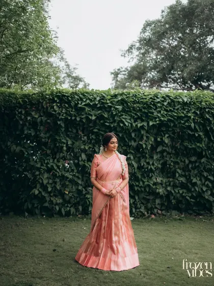 A full-length portrait of Dhriti in her elegant pink lehenga-saree, set against a wall of lush green ivy. The simplicity of the background makes her outfit stand out.
