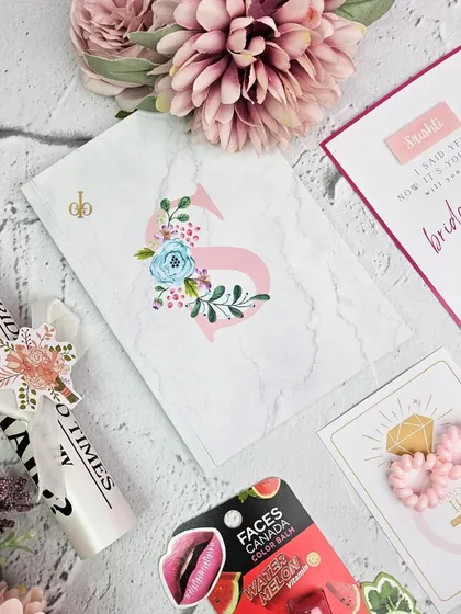 A close-up of the personalized items inside my bridesmaid hampers. This set includes a custom marble-print diary with a floral monogram, a "Bridesmaid Times" scroll, and other small, thoughtful goodies.