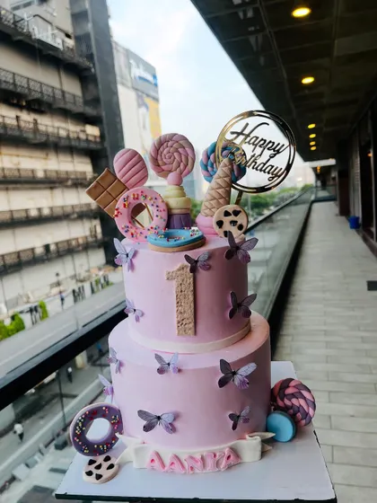 A delightful two-tier candyland themed cake for a first birthday. This eggless cake is adorned with fondant donuts, lollipops, an ice cream cone, and delicate butterflies, creating a sweet fantasy for a little one's celebration.