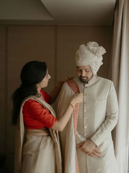 A moment between the groom and his mother as she helps him with his wedding attire, a classic ivory sherwani with a red-bordered stole.