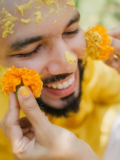 A close-up of the groom smiling as marigold flowers are playfully placed on his face during the haldi ceremony.