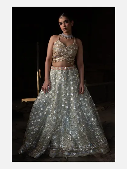 A studio-lit portrait of a model in a shimmering, light blue floral lehenga. The dark background makes the intricate silver and gold embroidery of the outfit stand out.