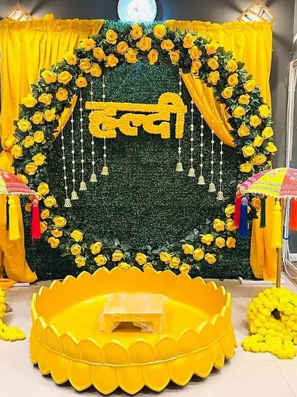 A beautiful Haldi ceremony setup featuring a lush green backdrop with a ring of yellow roses and the word 'Haldi' in Hindi. The decor is completed with traditional umbrellas and a central vessel for the ceremony.
