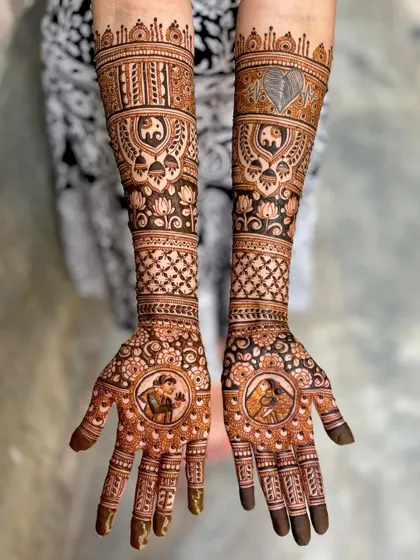 A beautiful bridal mehendi featuring portraits of the bride and groom in circular frames on the palms. The arms are decorated with elephant and lotus motifs.