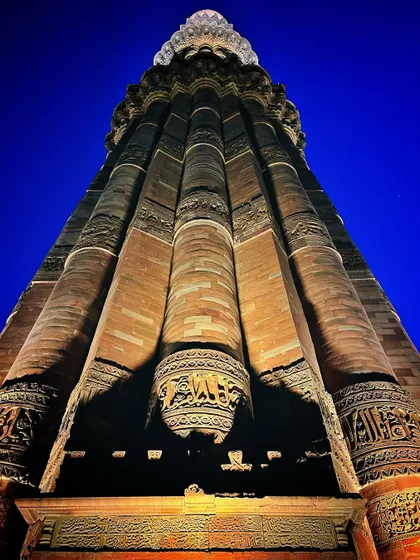 A dramatic low-angle shot of the Qutub Minar at night, emphasizing its fluted columns and detailed carvings against a deep blue sky.