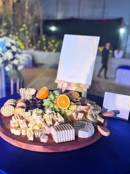 A sophisticated cheese platter set against a deep blue tablecloth at a Godrej Properties event. The board features a mix of cheeses, crackers, and fruits, with a custom menu card displayed alongside.