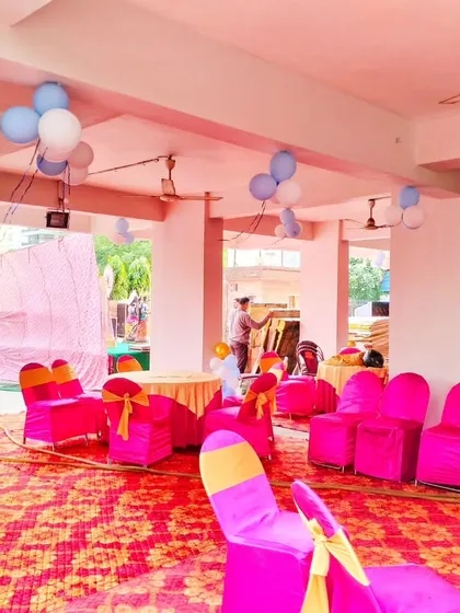 A view of the seating arrangement for a Kuan Pujan celebration. The space is decorated with balloon clusters on the ceiling and pillars, with bright pink chair covers adding to the festive look.