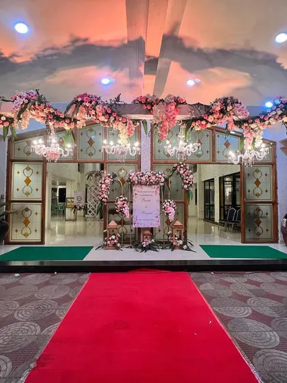 A grand entrance for a reception, featuring a floral archway in shades of pink, leading to a red carpet welcome. Chandeliers add a touch of classic elegance.