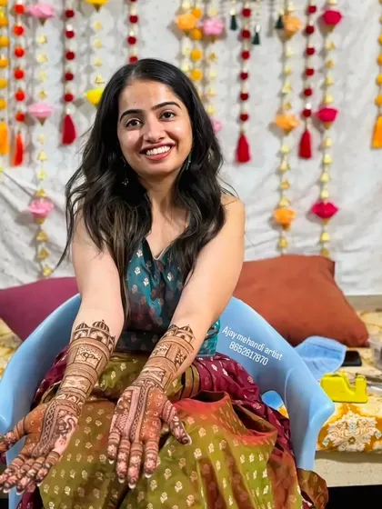 A smiling bride seated in front of a colorful mehndi backdrop, her hands beautifully decorated with my art.