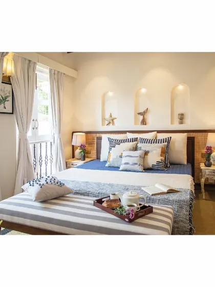 This bedroom in a Goan holiday home uses a cool color palette of blues and whites. We designed the bed with a wood and cane headboard, and added arched niches in the wall for displaying small decor items, adding a touch of local architectural charm.