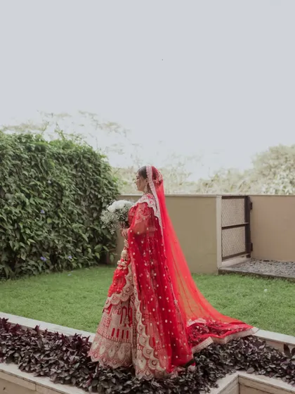 A side profile of the bride walking in the garden, her long red veil trailing behind her.